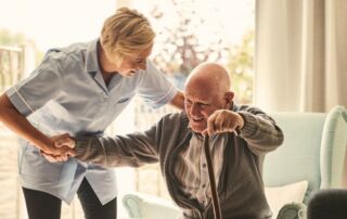 In-home caregiver assists elderly man with cane out of his chair while providing home care services