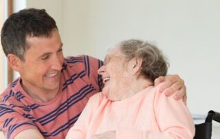 Young man has his arm over the shoulder of an elderly woman in a wheelchair as she laughs